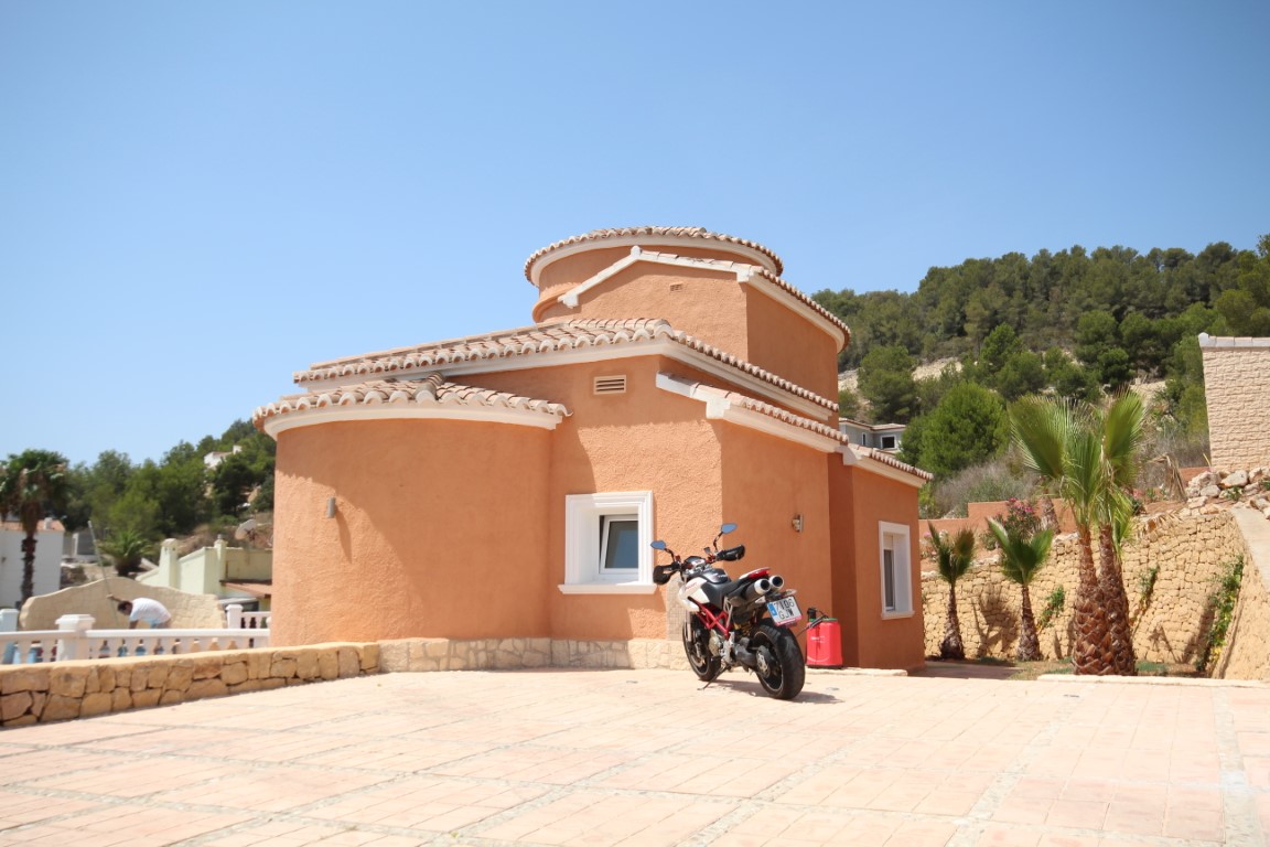 Mediterrane Fassade mit geschwungenen terracottafarbenen Wänden, großzügigem gepflastertem Parkplatz und Natursteinmauern mit Palmen.