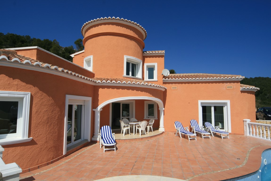 Mediterrane Villa mit terracottafarbener Fassade, markantem Rundturm, weitläufiger Fliesenterrasse mit gestreiften Liegen und einem schattigen Bogengang.