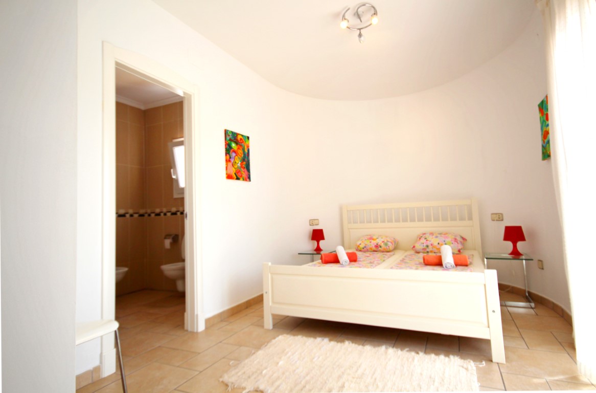 Schlafzimmer mit geschwungener Wand, weißem Holzbettrahmen und direktem Zugang zu einem beige gefliesten En-Suite-Badezimmer.