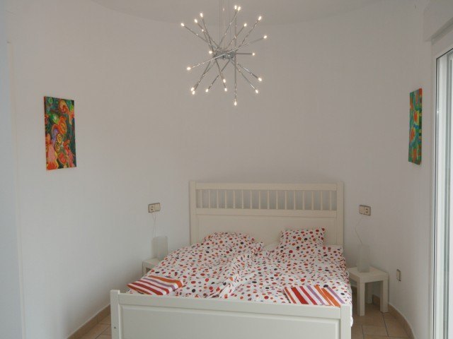 Schlafzimmer mit weißem Holzbett, moderner Deckenleuchte im Sterndesign und direktem Zugang zum Außenbereich durch Glastüren.