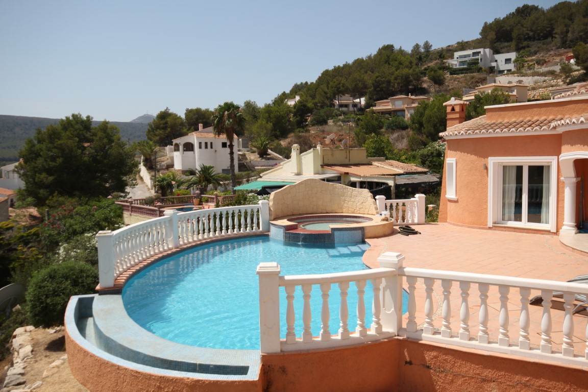 Sonnenterrasse mit einem geschwungenen Infinity-Pool, integriertem Whirlpool mit Natursteinwand und klassischer weißer Balustrade vor der Villa.