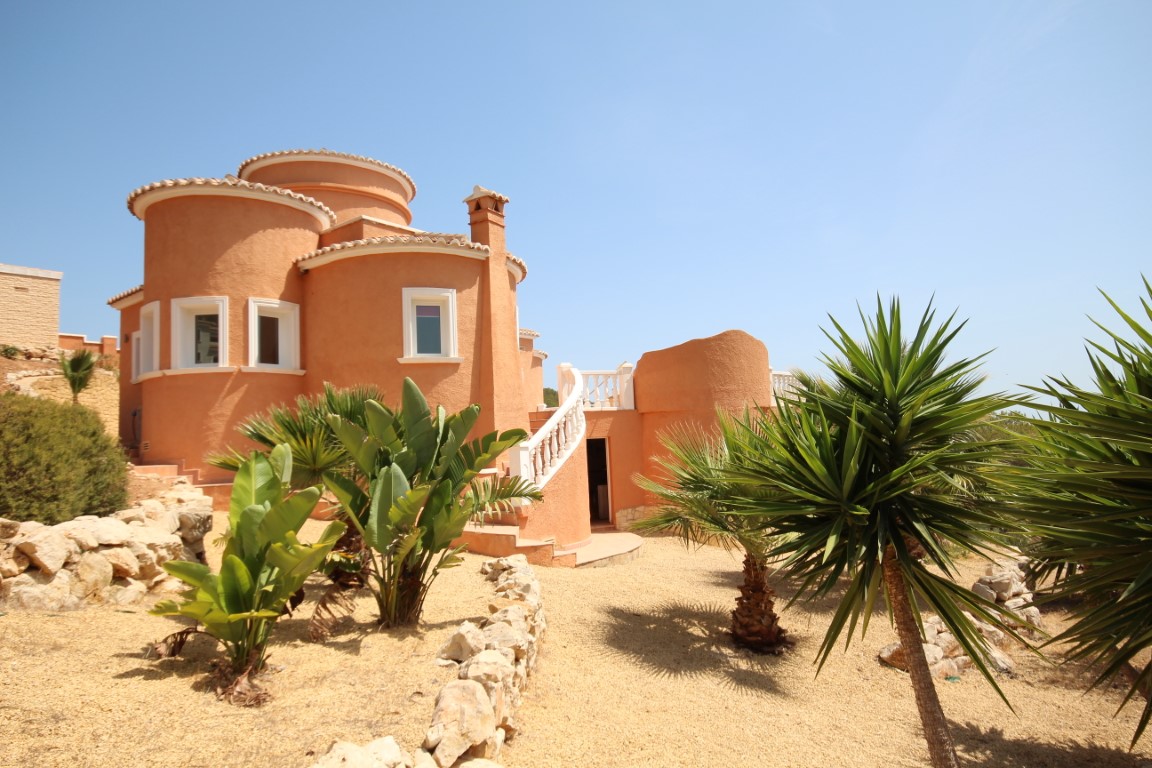 Terracottafarbene Villa mit runden architektonischen Formen, einer weißen Außentreppe mit Balustrade und einem mediterranen Kiesgarten mit Palmen.