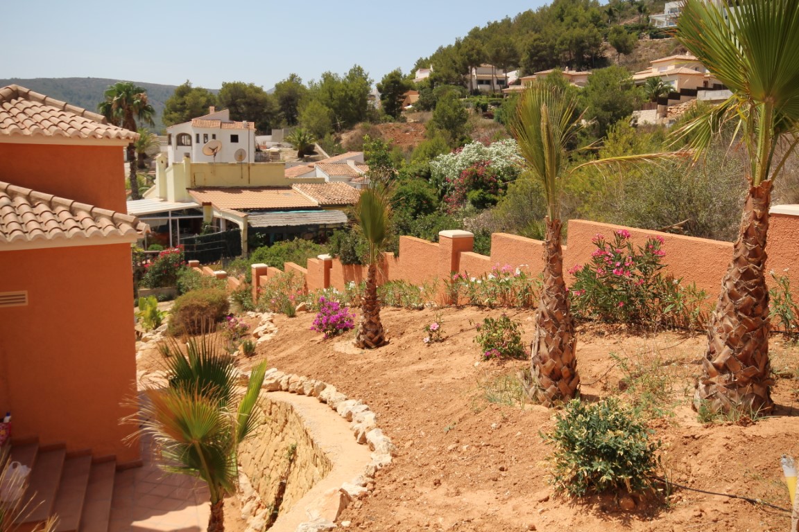 Terrassierter mediterraner Garten mit jungen Palmen, blühenden Sträuchern und terrakottafarbenen Mauern mit Blick auf die umliegende Hügellandschaft.