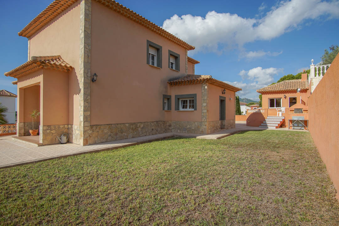 Außenansicht der Villa mit erdfarbener Fassade, Natursteinakzenten und Rasen. Im Hintergrund ist die separate Außenküche mit Holzofen zu sehen.