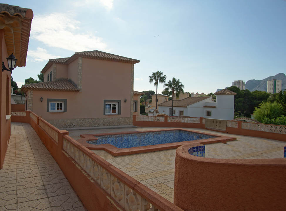 Außenansicht des mit blauen Mosaikfliesen gestalteten Pools und der großzügigen Pflasterterrasse, umgeben von terracottafarbenen Mauern mit Natursteinakzenten.