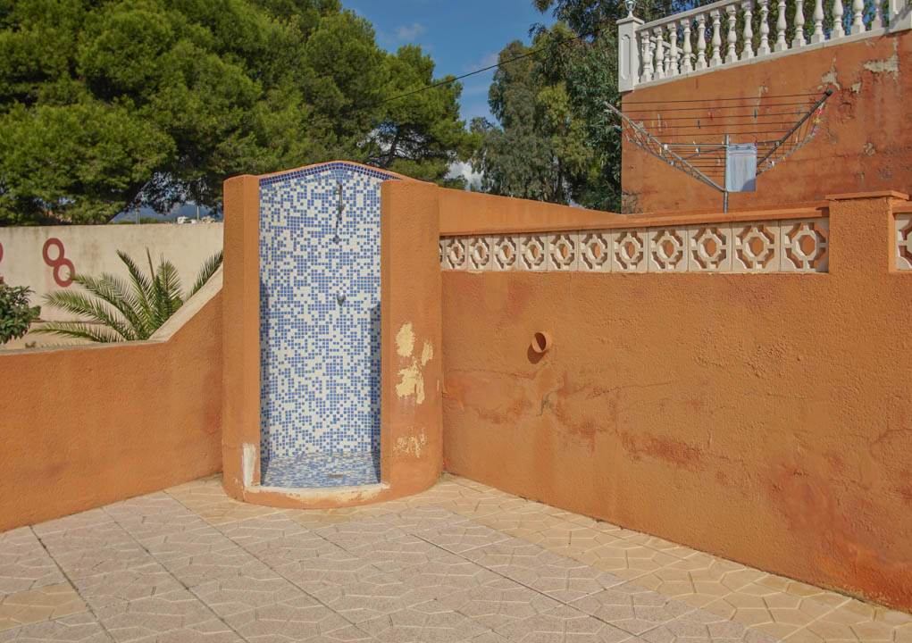 Außendusche mit blau-weißer Mosaikverkleidung, eingefasst von terrakottafarbenen Mauern mit dekorativen Formsteinen und gemustertem Steinboden.