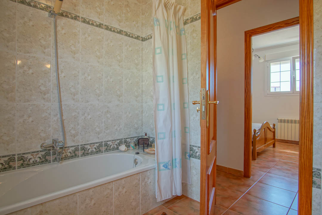 Badezimmer mit Einbauwanne, dekorativen Fliesenelementen und Massivholz-Türrahmen, die in ein helles Schlafzimmer führen.