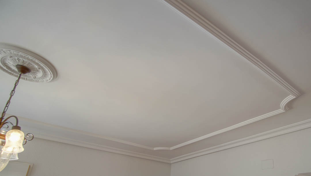 Detail der Decke mit klassischen Zierleisten und einer zentralen Deckenrosette. Die Beleuchtung besteht aus einem traditionellen Kronleuchter mit Glasschirmen.