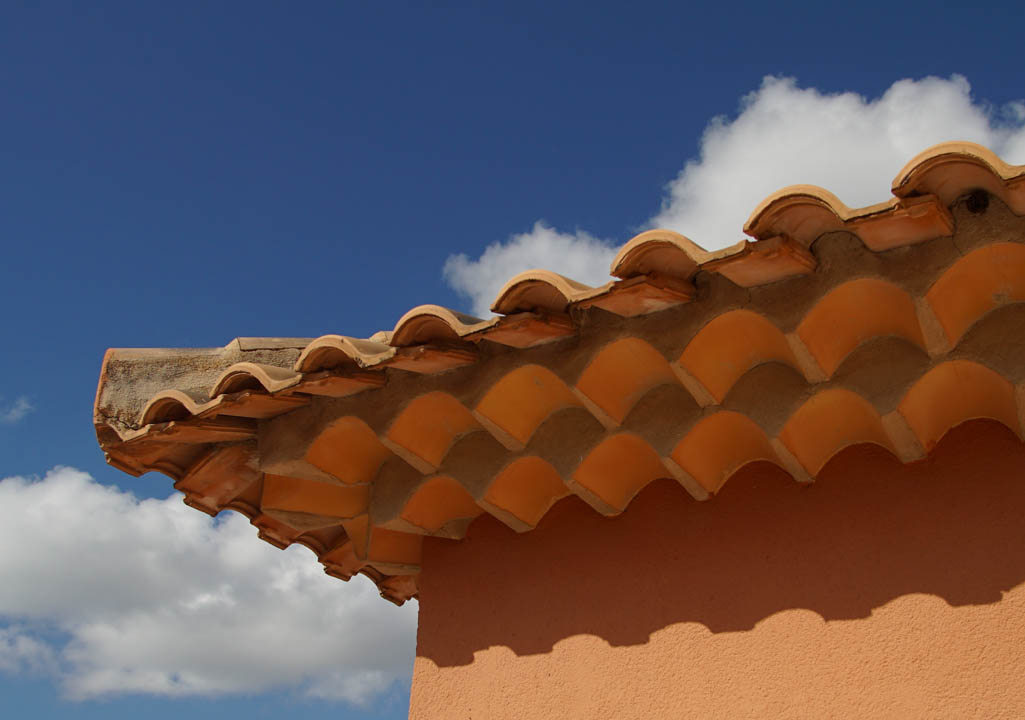 Detailansicht der traditionellen Tondachziegel mit dekorativem, dreilagigem Gesims an einer terrakottafarbenen Außenwand unter blauem Himmel.