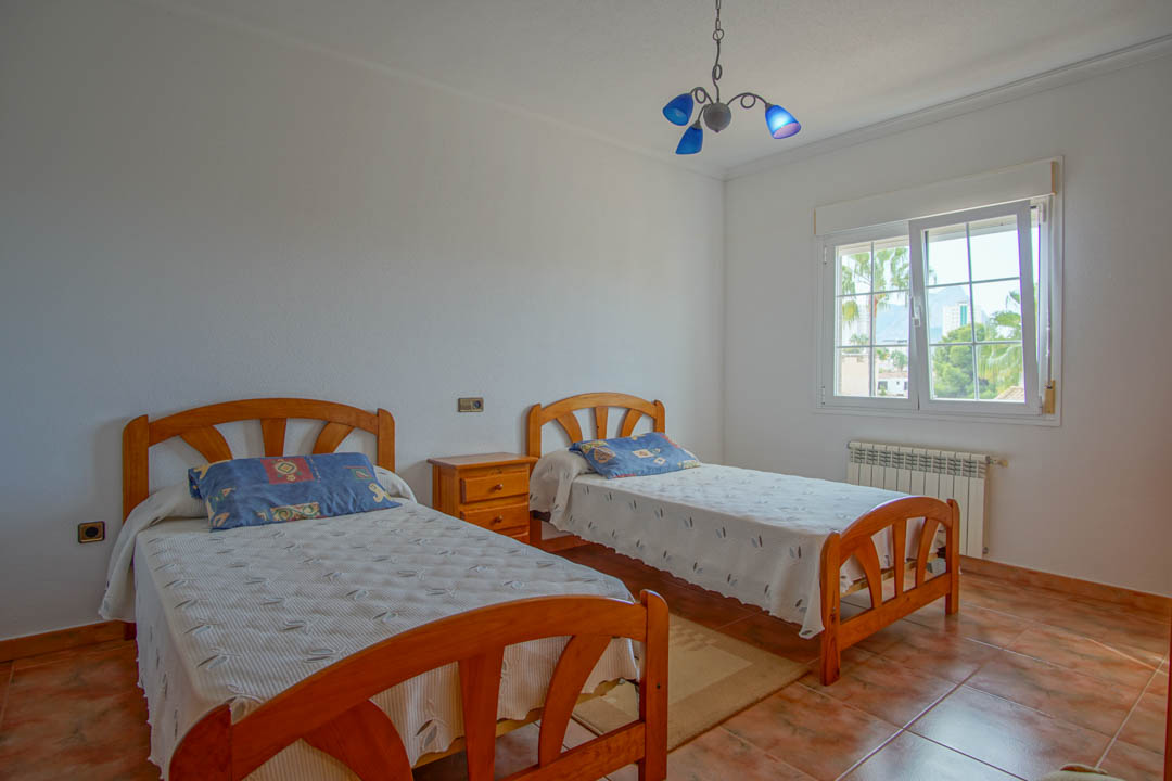 Helles Schlafzimmer mit zwei Massivholz-Einzelbetten, Keramikfliesen und Fenster mit Blick auf die umliegende Landschaft und Palmen.