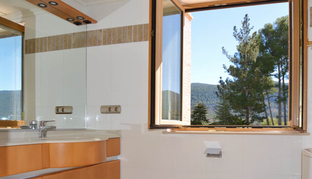 Badezimmer mit Holzwaschtisch, großem Spiegel und offenem Fenster mit Blick auf die umliegenden Pinienwälder und die Berglandschaft der Finca.