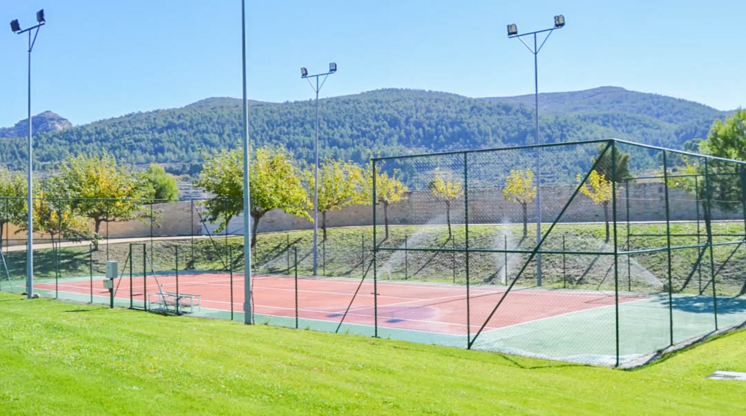 Eingezäunter Tennisplatz mit roter Spielfläche und Flutlichtanlage, eingebettet in eine gepflegte Grünanlage mit Blick auf die umliegende Berglandschaft.