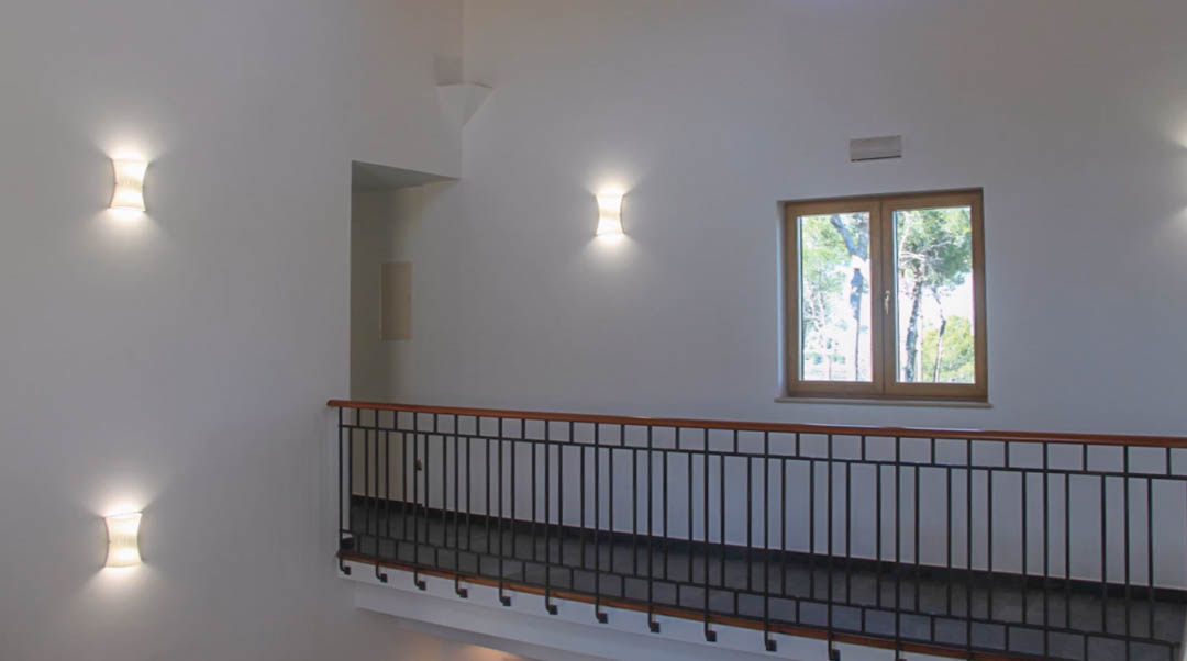 Flur im Obergeschoss mit schwarzem Metallgeländer, Handlauf aus Holz, Wandleuchten und einem Holzfenster mit Blick auf die umliegende Baumlandschaft.