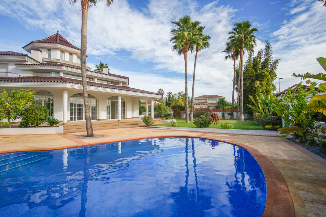 Außenansicht der Kolonialvilla mit geschwungenem Pool, gewachsenem Palmengarten und weißer Fassade mit Rundbogenfenstern und Säulen.
