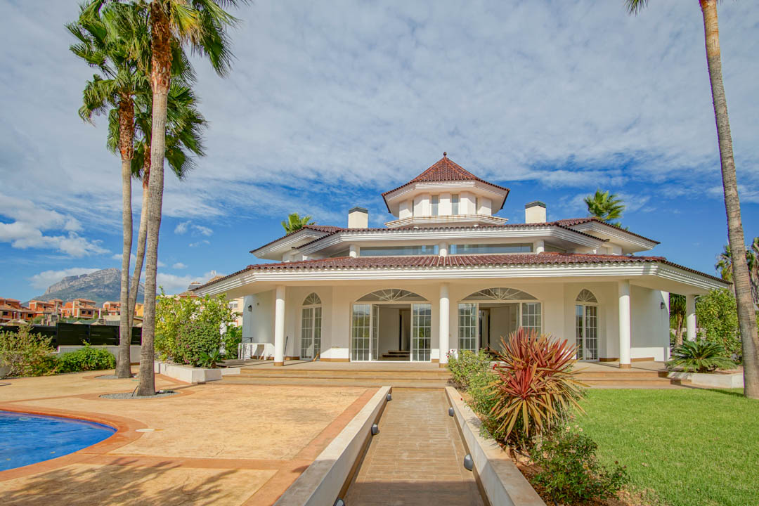Außenansicht einer Kolonialvilla mit weißer Fassade, Säulenportikus, zentralem achteckigem Turm und Garten mit Palmen und Pool.