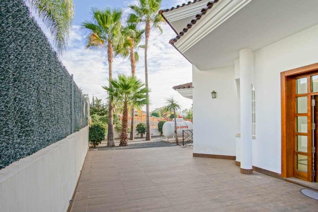 Außenansicht mit gefliester Terrasse, weißer Fassade, einer runden Säule und dem Gartenbereich mit hohen Palmen hinter dem Sichtschutzzaun.