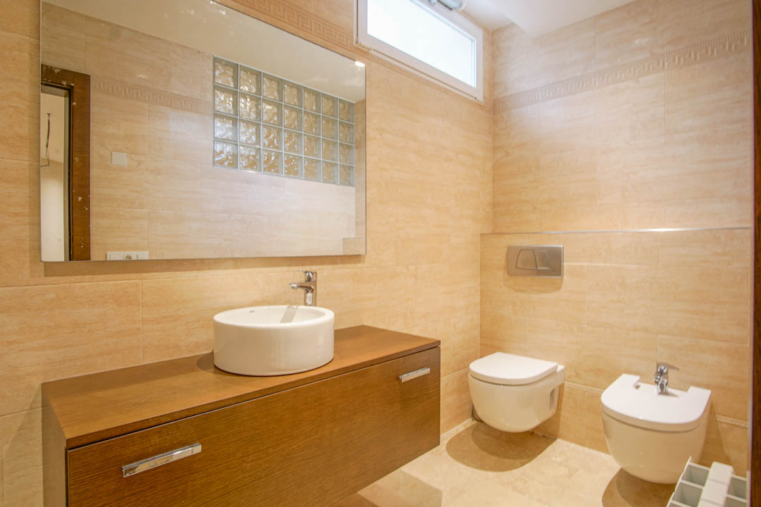 Badezimmer mit beigen Fliesen, schwebendem Holzwaschtisch, Aufsatzwaschbecken, wandhängenden Sanitäranlagen und Spiegel mit Reflexion einer Glasbausteinwand.