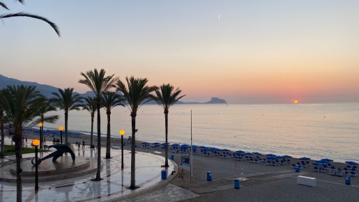 Blick auf den Sonnenaufgang von der nahegelegenen Promenade mit Palmen, einer Ankerskulptur und dem Sandstrand mit blauen Sonnenschirmen am Mittelmeer.