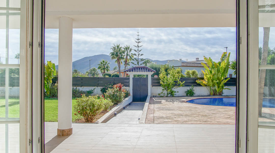 Blick aus dem Innenraum auf die überdachte Terrasse und den Garten mit Pool, Palmen und Bergblick, eingerahmt von weißen Sprossenfenstertüren.
