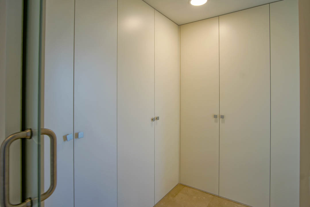 Deckenhohe Einbauschränke in Weiß mit quadratischen Metallgriffen, Natursteinboden und Beleuchtung durch Deckeneinbaustrahler.