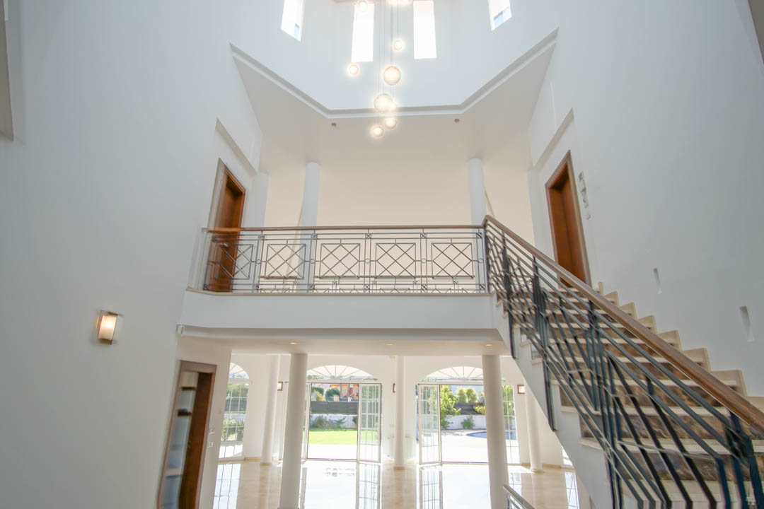 Doppelt hohe Eingangshalle mit Marmortreppe, schmiedeeisernem Geländer und oberer Galerie, die durch moderne Pendelleuchten und große Rundbogenfenster erhellt wird.