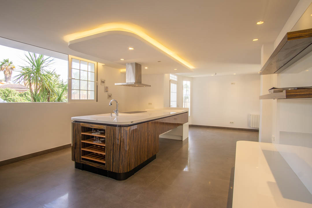 Moderne Küche mit einer Kochinsel in Holzoptik, weißer Arbeitsplatte und Dunstabzugshaube aus Edelstahl. Die indirekte LED-Deckenbeleuchtung schafft ein elegantes Ambiente.
