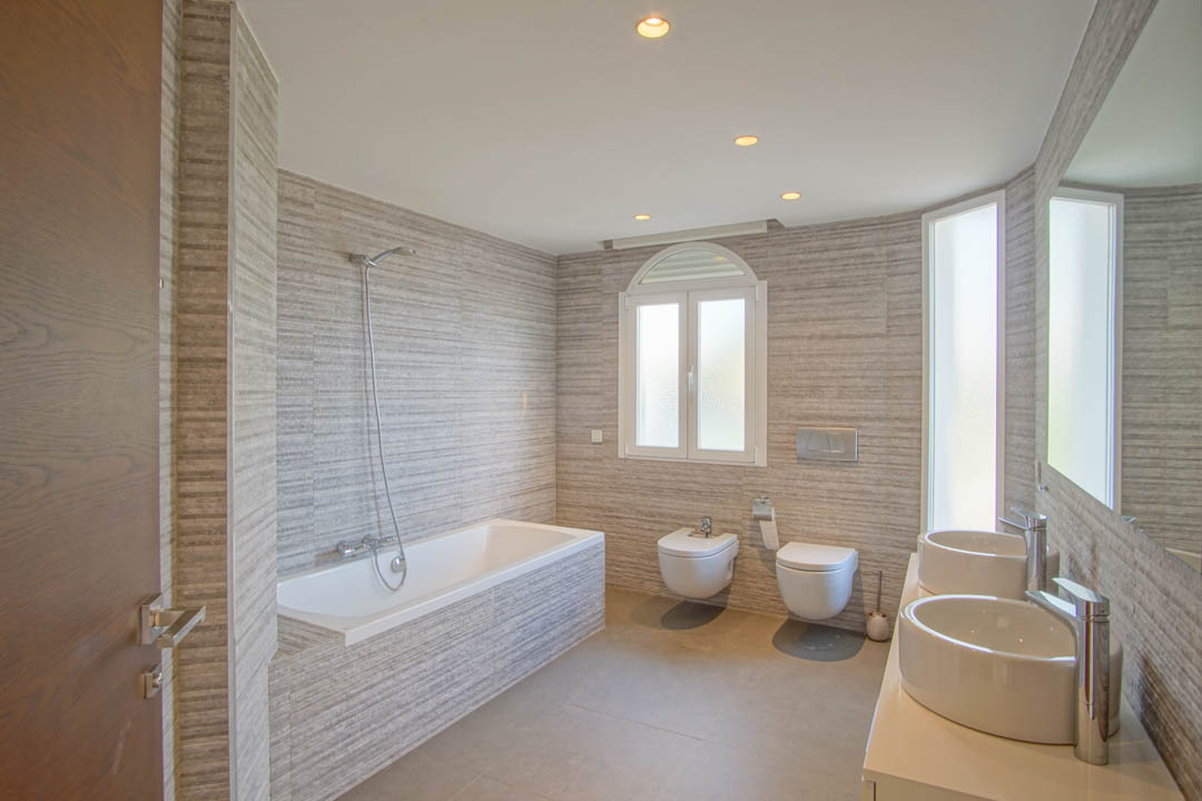 Modernes Badezimmer mit Doppel-Aufsatzwaschbecken, Einbaubadewanne und strukturierten Wandfliesen. Ausgestattet mit wandhängendem WC und natürlichem Licht durch ein Rundbogenfenster.