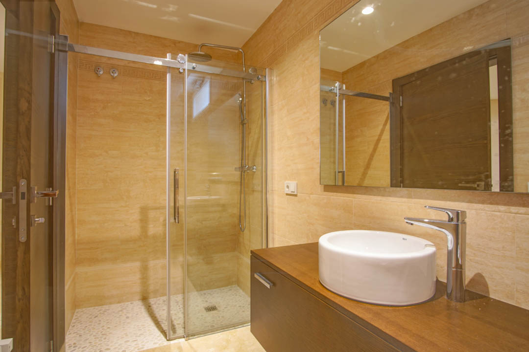Modernes Badezimmer mit Glasdusche, Kieselsteinboden, rundem Aufsatzwaschbecken auf dunklem Holzwaschtisch und warmen beigen Wandfliesen.