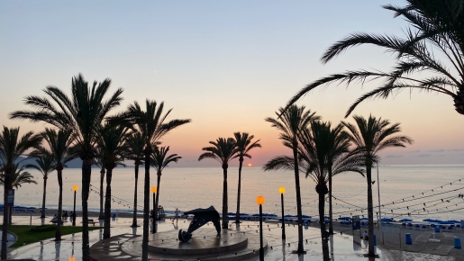 Panoramablick auf den Sonnenaufgang über der Küstenpromenade mit hohen Palmen, stimmungsvoller Beleuchtung und einer Skulptur direkt am Mittelmeer.