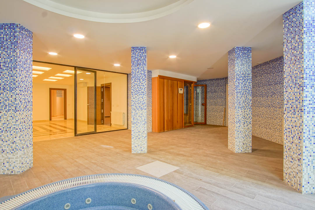 Spa-Bereich mit kreisförmigem Whirlpool, Holzsauna und blau gemusterten Mosaiksäulen. Ausgestattet mit Deckeneinbaustrahlern und Glaswänden zu den weiteren Kellerräumen.