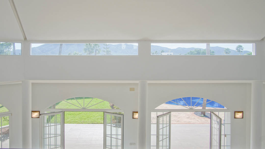 Wohnbereich mit doppelter Deckenhöhe, Rundbogentüren zum Garten und Pool sowie Oberlichtern mit weitem Blick auf die umliegenden Berge und Palmen.