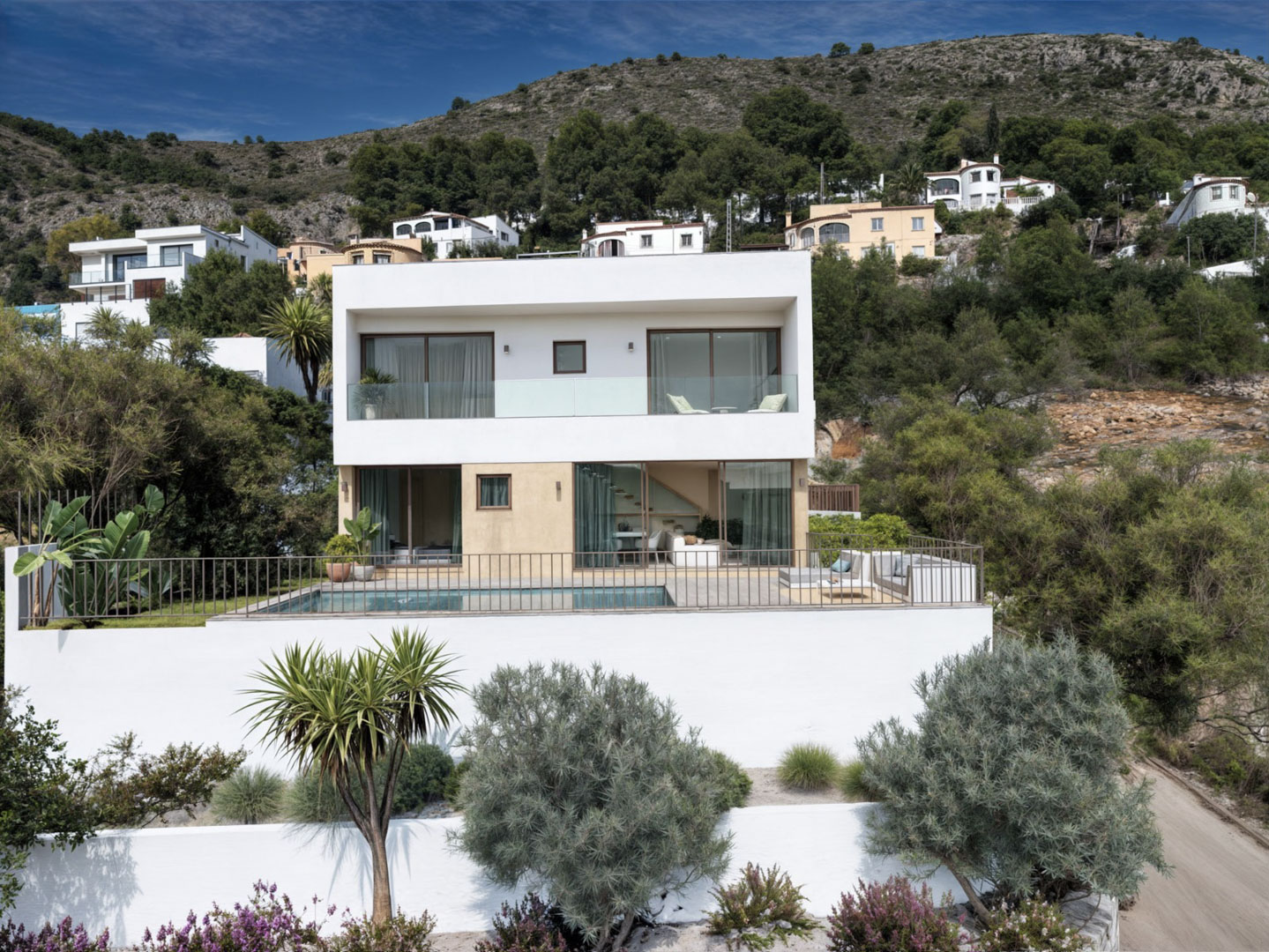 Außenansicht der modernen Villa mit weiß-beiger Fassade, privatem Pool, großen Glasschiebetüren und Balkonen mit Glasgeländer vor einer bergigen Kulisse.