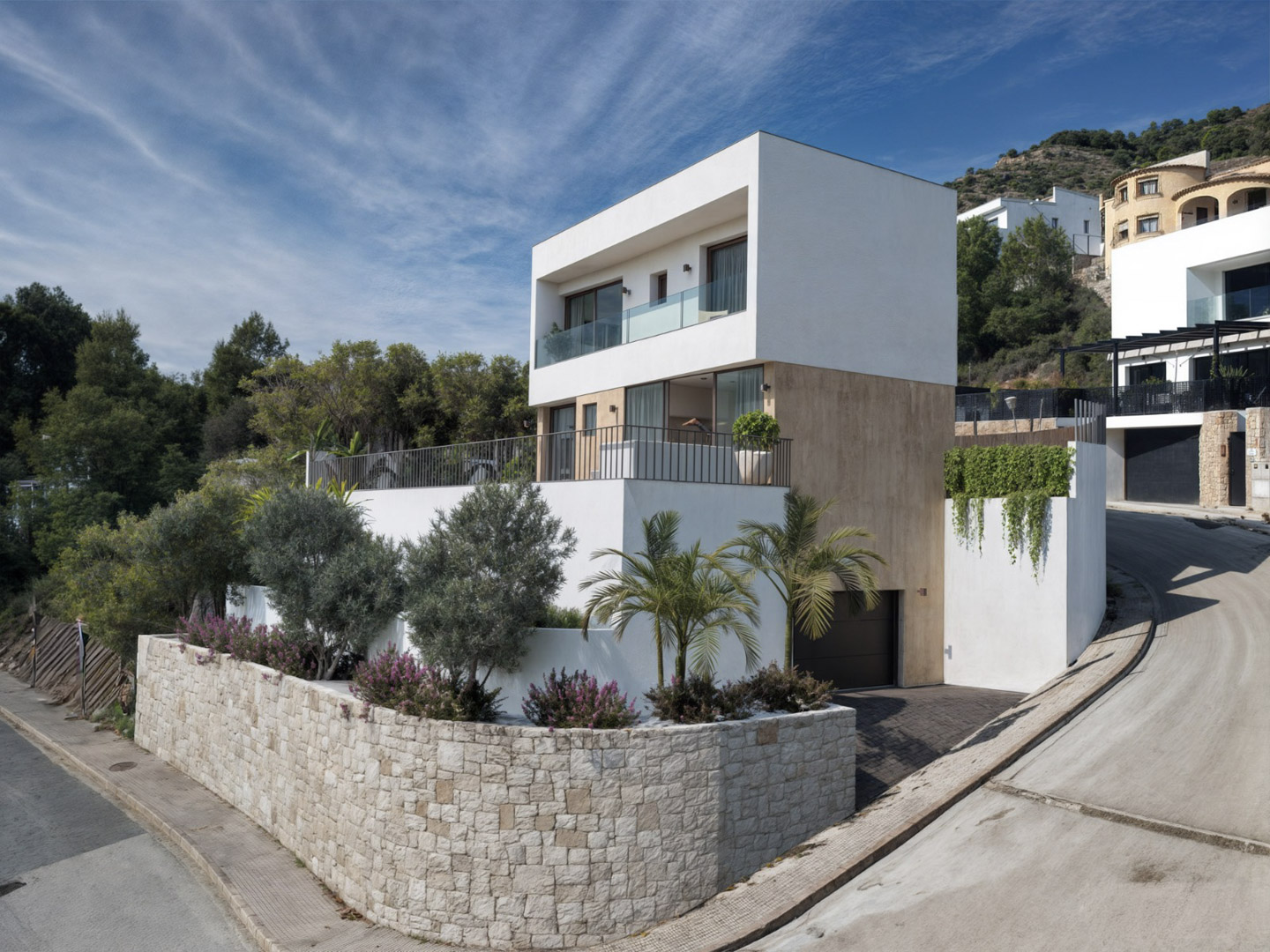Moderne dreistöckige Villa mit Natursteinfassade, Terrassen mit Glasgeländern und einer im Souterrain integrierten Garage in Hanglage.