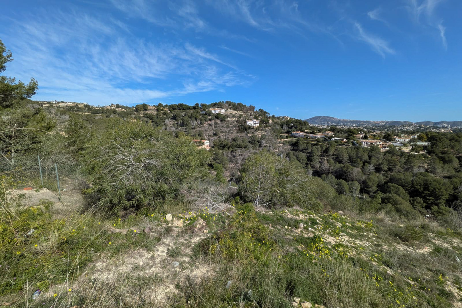 Erhöhter Blick vom steilen Grundstück auf die natürliche Vegetation und die bewaldeten Hügel von Benissa unter strahlend blauem Himmel.