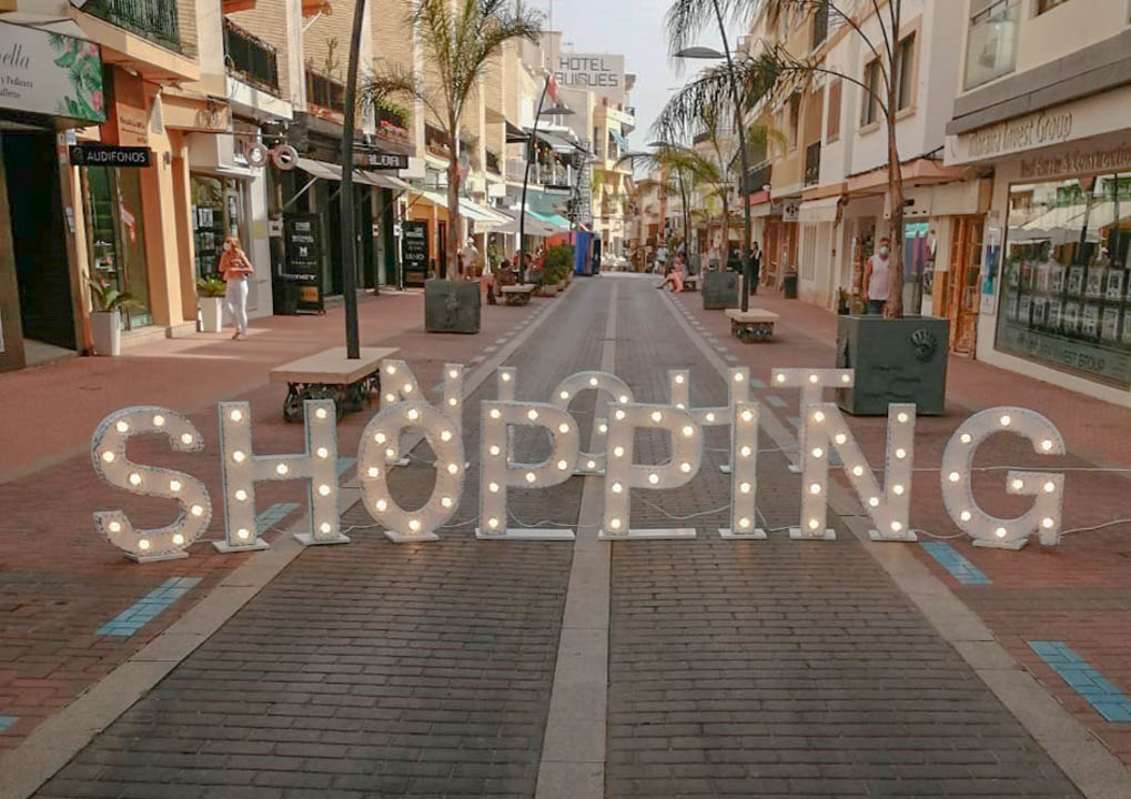 Fußgängerzone im Zentrum von Moraira mit Palmen, lokalen Geschäften und einem großen, beleuchteten Schriftzug auf dem gepflasterten Weg.