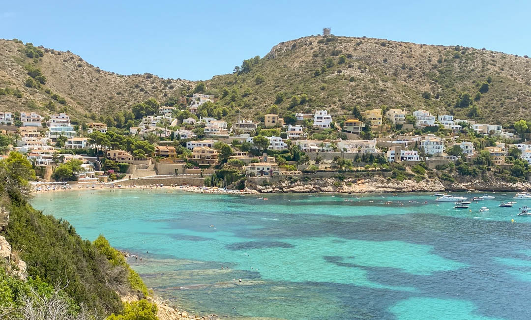 Panoramablick auf die Bucht von El Portet in Moraira mit türkisblauem Wasser, vor Anker liegenden Booten und weißen Villen an den mediterranen Hängen.