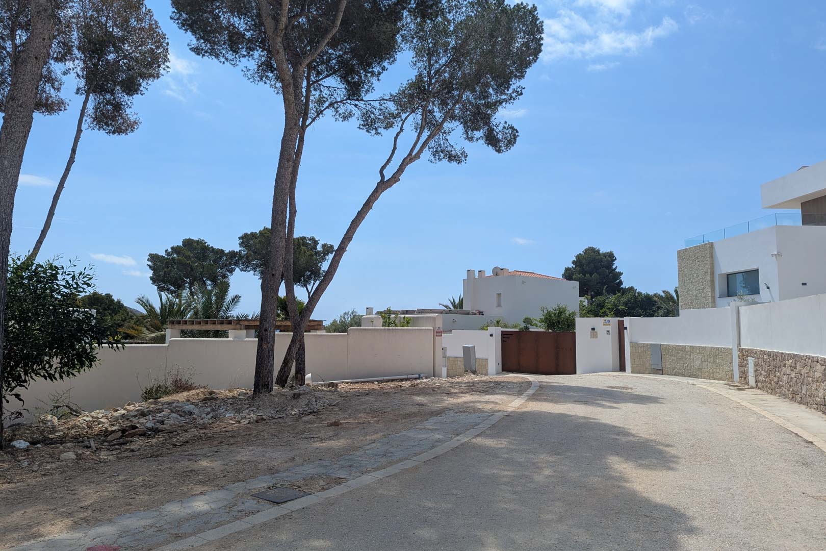Zufahrt zum Grundstück in einem Wohngebiet von Moraira mit weißen Umfassungsmauern, alten Pinien und einer gepflasterten Straße unter blauem Himmel.