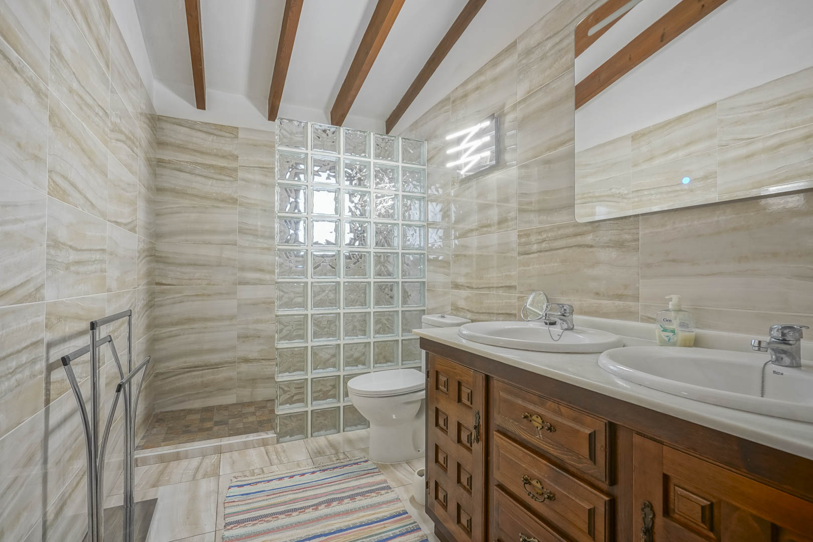 Badezimmer mit Glasbaustein-Duschwand, Waschtisch aus Holz mit Doppelwaschbecken und geäderten Fliesen.