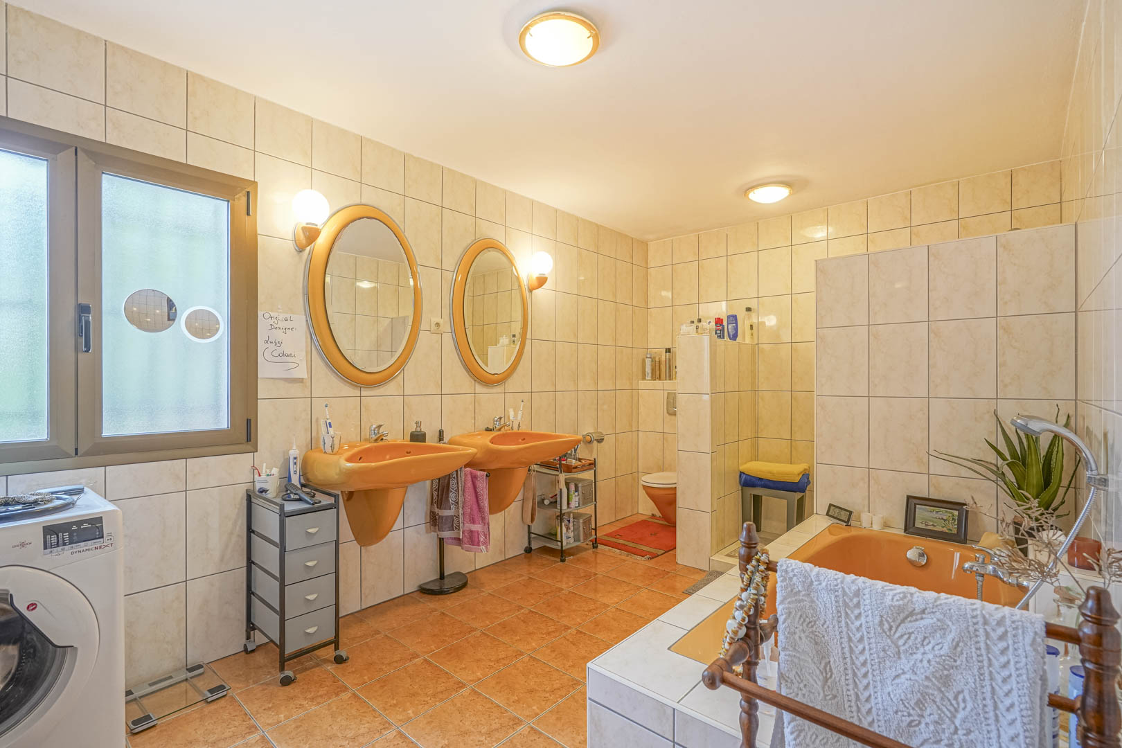Badezimmer mit orangefarbenen Waschbecken und passender Badewanne, cremefarbenen Fliesen und Fenster mit Tageslicht.