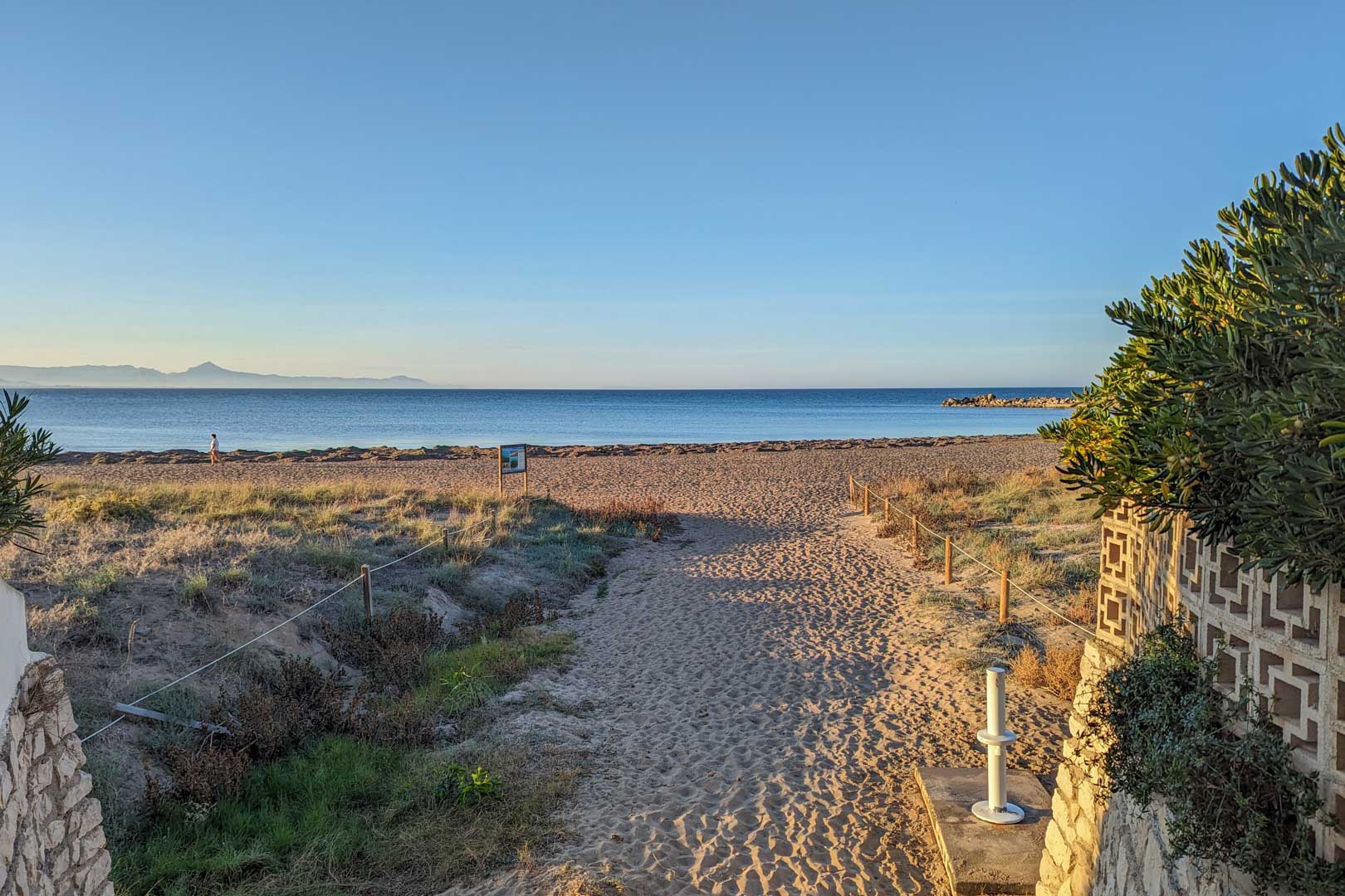 Blick auf Sandstrand mit abgegrenztem Weg, Küstenvegetation und blauem Meer unter klarem Himmel.