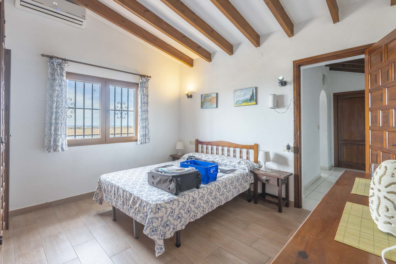 Helles Schlafzimmer mit Holzbalken, Doppelbett mit gemusterter Bettwäsche und Blick nach draußen.
