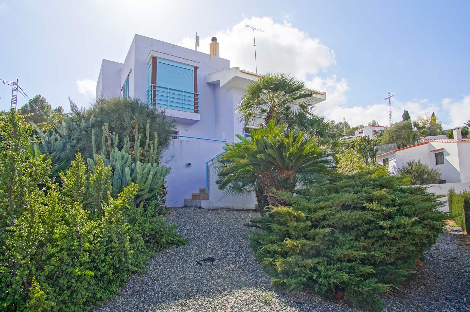 Außenansicht der lavendelfarbenen Fassade, eingebettet in einen mediterranen Garten mit Kakteen, Palmen und Sträuchern auf Kiesboden.