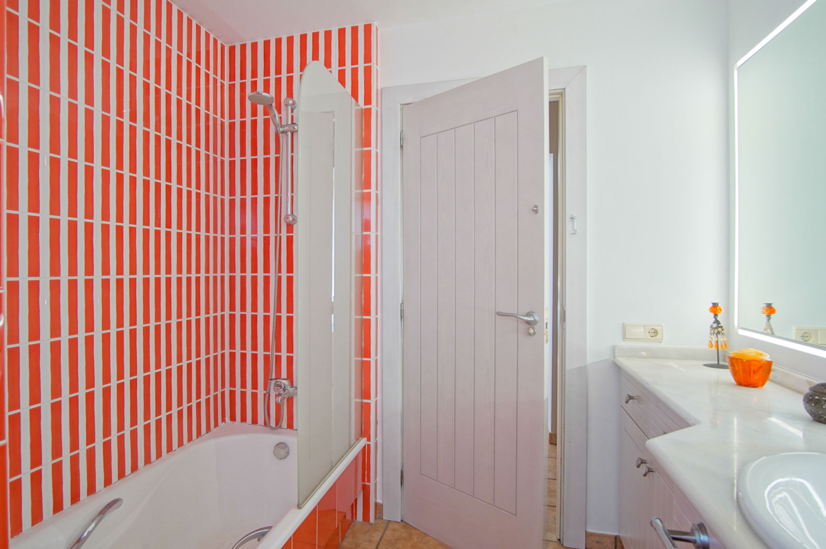 Badezimmer mit Badewanne und Glaswand, akzentuiert durch orange-weiße Vertikalfliesen, einen weißen Waschtisch mit Marmorplatte und einen hinterleuchteten Spiegel.