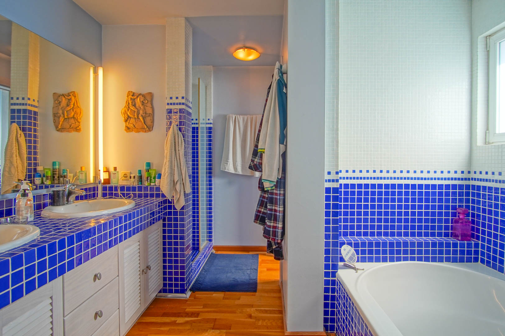Badezimmer mit blau-weißem Mosaik-Waschtisch, Doppelwaschbecken, Holzboden und Einbaubadewanne. Warme Beleuchtung und weiße Lamellentüren prägen das funktionale Design.