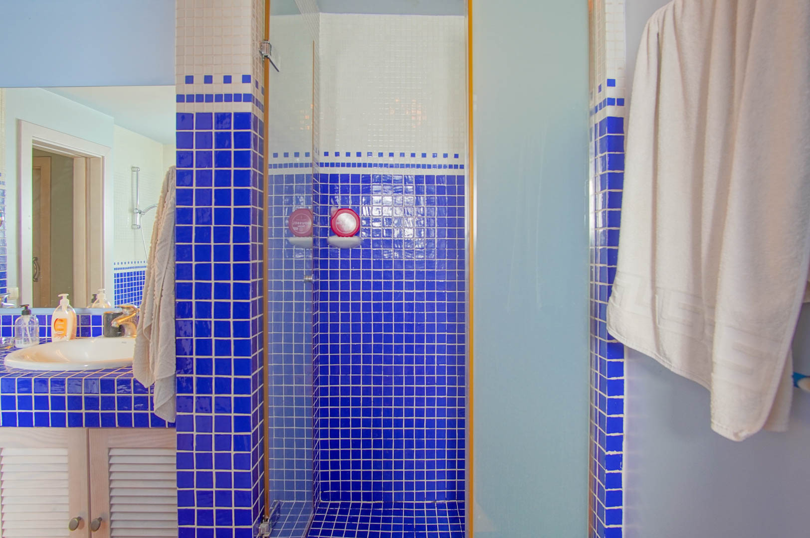 Badezimmer mit blau-weißen Mosaikfliesen, einer begehbaren Dusche mit Glastür und einem Waschtisch mit Lamellentüren aus Holz.