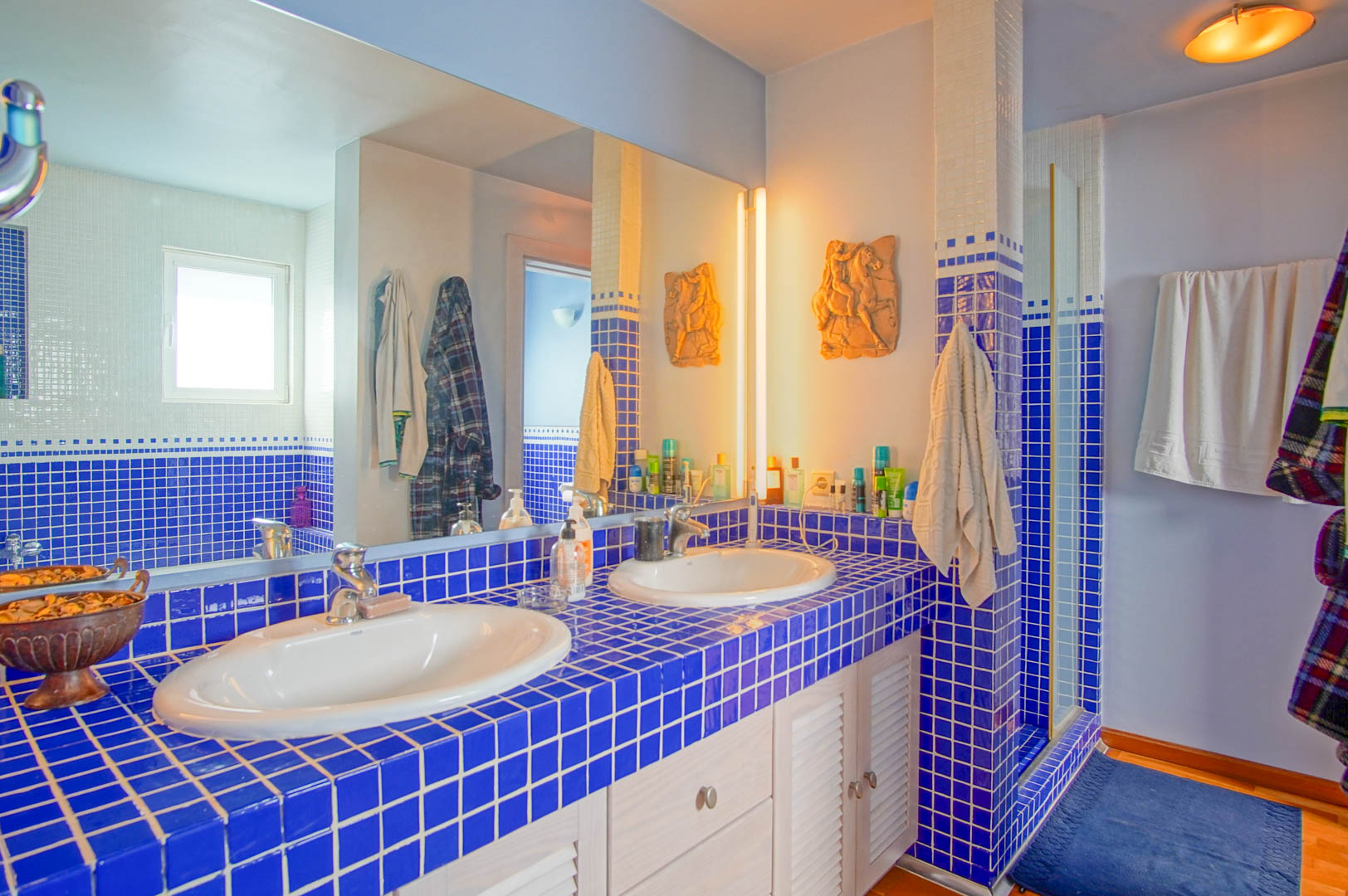Badezimmer mit Doppelwaschbecken auf blauen Fliesen und weißen Lamellenschränken. Ein großer Wandspiegel reflektiert den Duschbereich und das warme Licht.