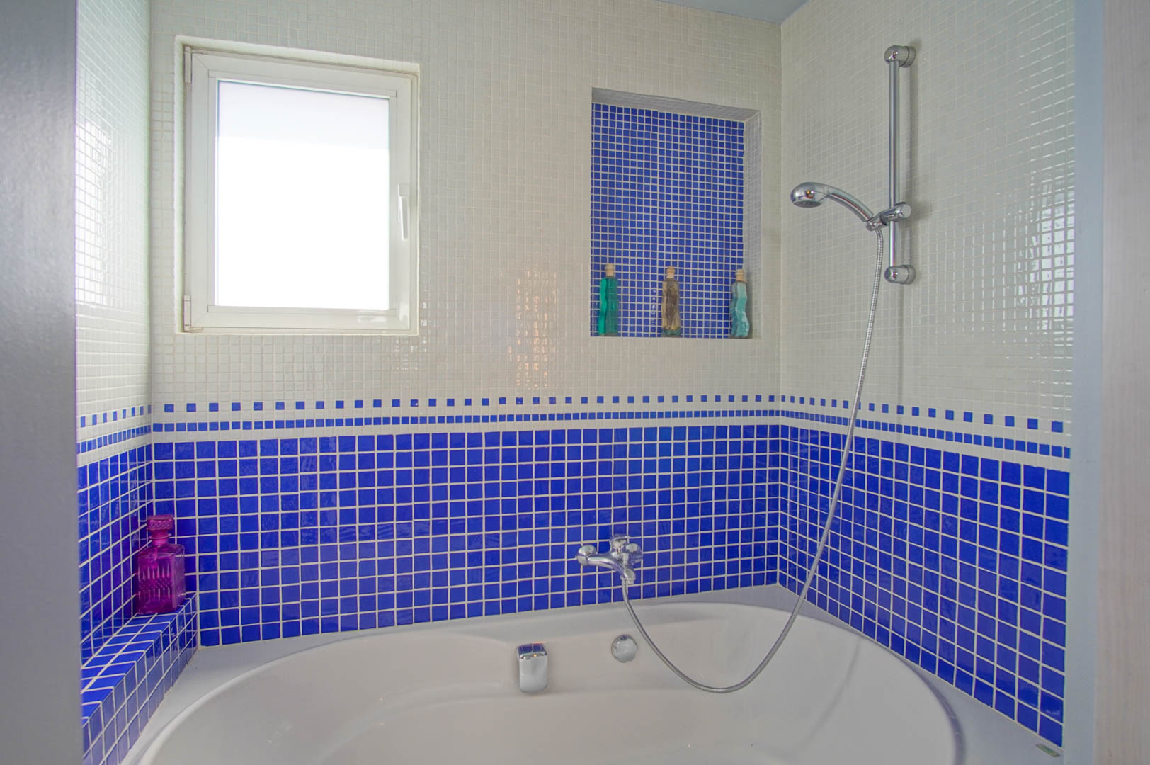 Badezimmer mit ovaler Badewanne und weiß-blauen Mosaikfliesen. Ausgestattet mit einer integrierten Wandnische für Utensilien und einem Fenster für Tageslicht.