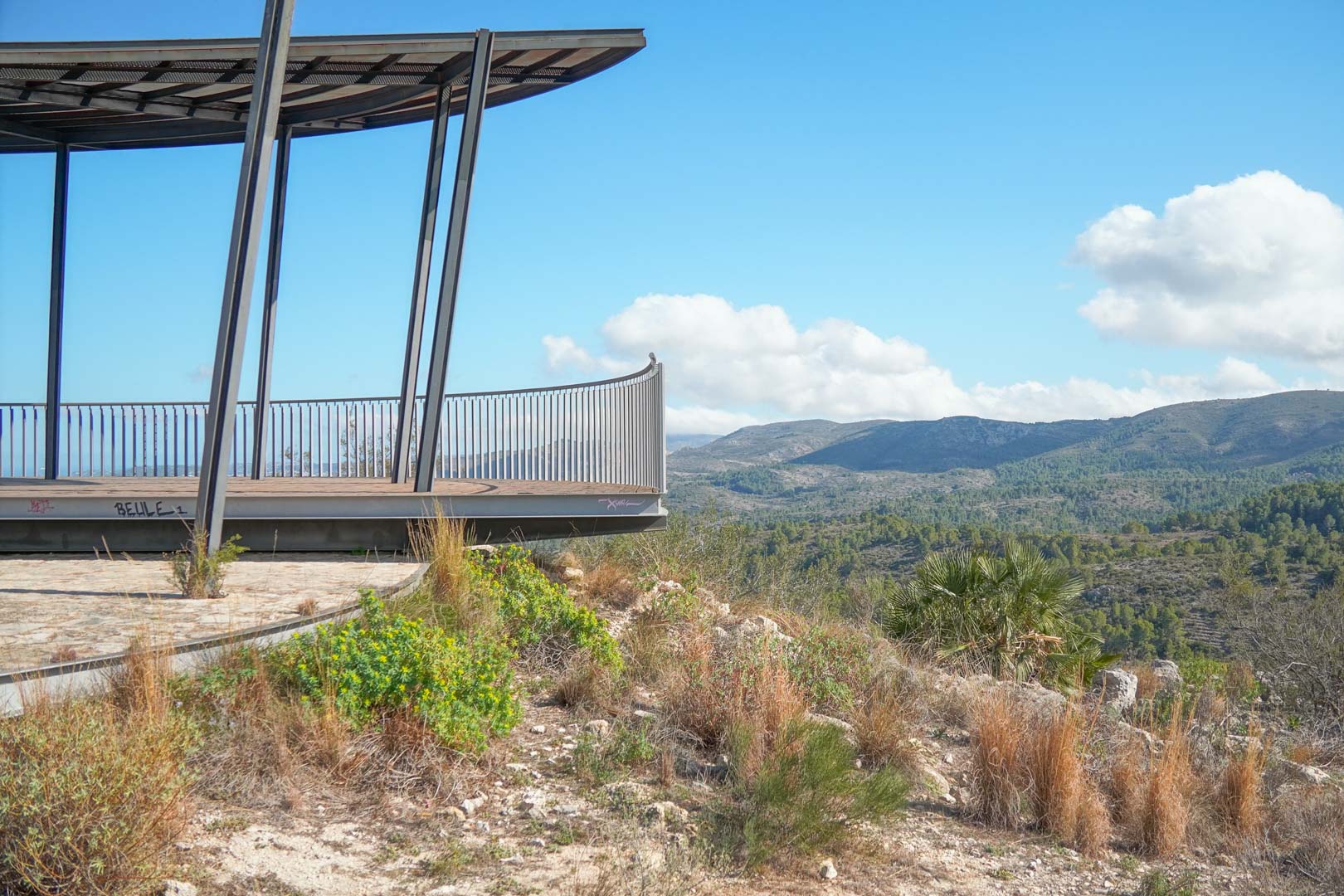 Erhöhte Aussichtsplattform mit Stahlrahmen und Metallgeländer, die einen weiten Panoramablick auf die Gebirgskette und das mediterrane Tal bietet.