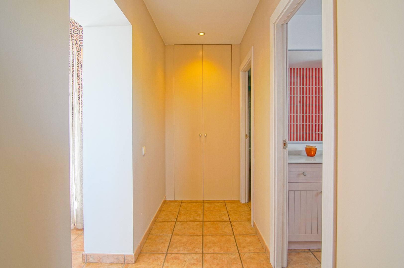 Flur mit Terrakotta-Fliesen, einem raumhohen Einbauschrank und Blick in ein Badezimmer mit markanten rot-weiß gestreiften Wandfliesen und weißem Waschtisch.