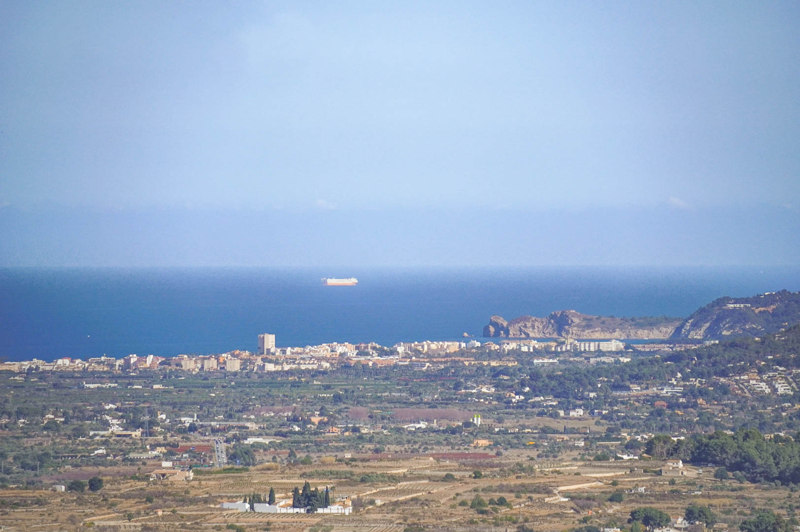 Freier Panoramablick auf die Mittelmeerküste mit der Silhouette der Küstenstadt und dem Meer, an dessen Horizont Schiffe zu sehen sind.